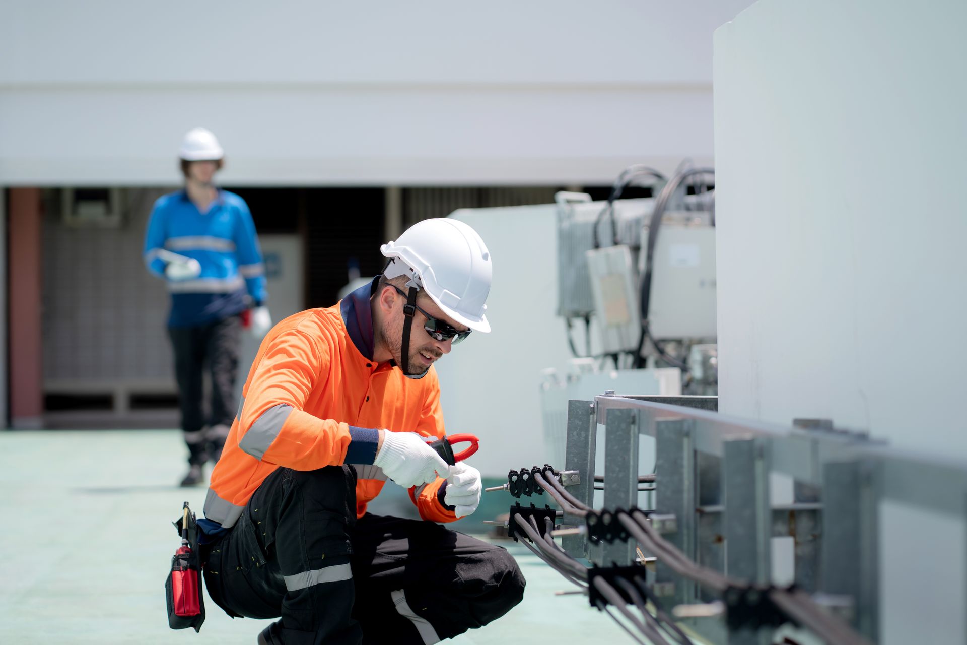Two technicians in hard hats and high-visibility gear work on electrical equipment outdoors.