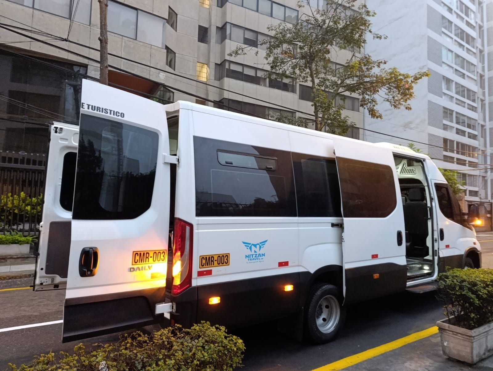 Furgoneta blanca de transporte público estacionada en una calle de la ciudad junto a edificios de apartamentos, con una puerta lateral y una puerta trasera abiertas.