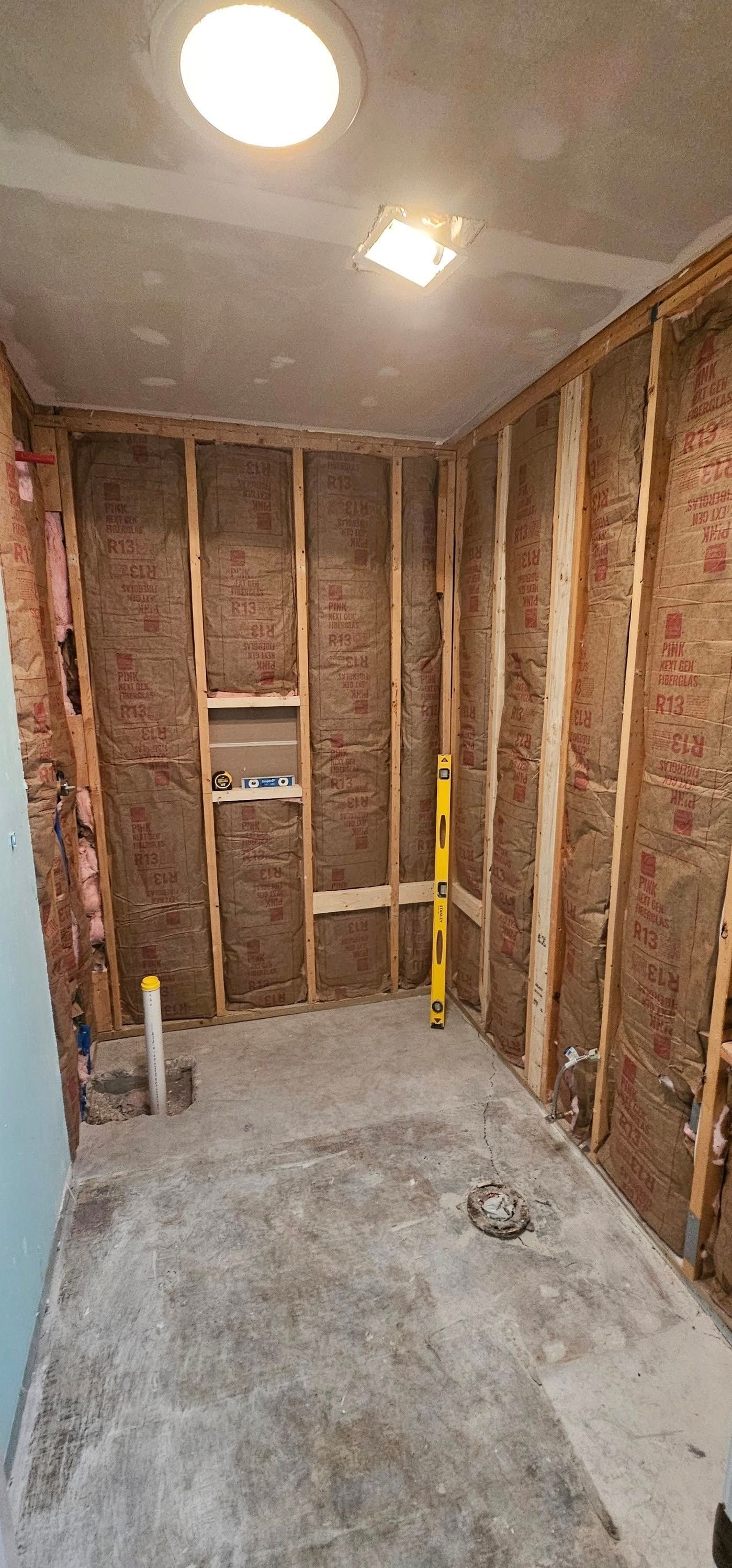 A room undergoing renovation with exposed wooden wall studs, insulation, plumbing fixtures, and a level leaning against wall.