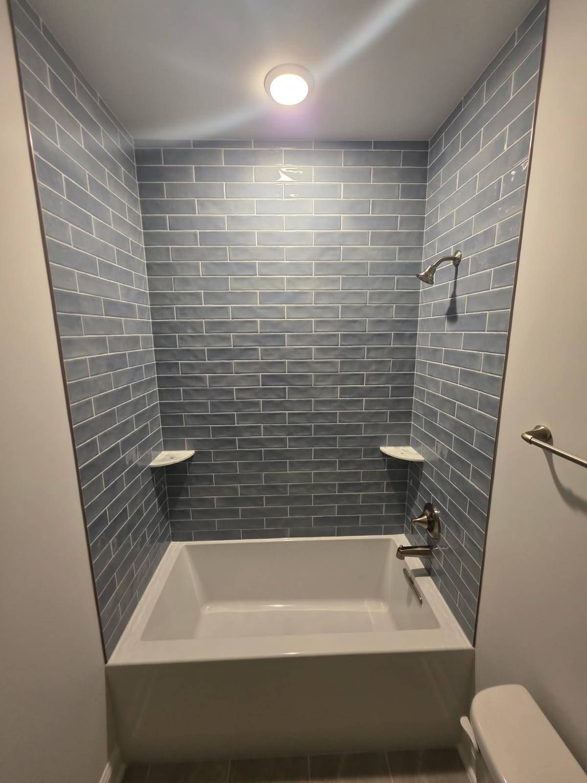A bathroom tub alcove with blue brick-patterned wall tiles, white shelves, and a silver showerhead and faucet.