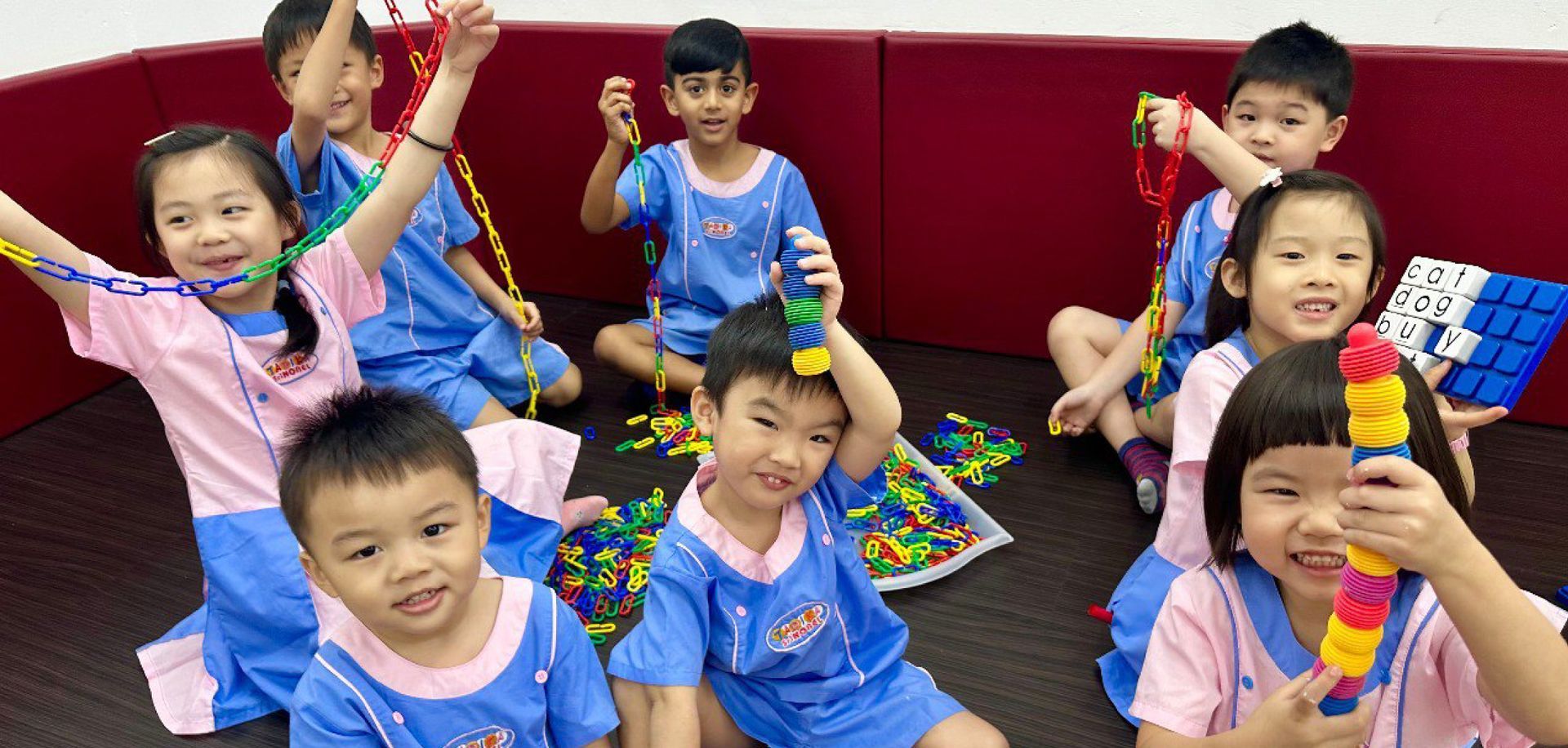 Children in blue uniforms and pink shirts play with colorful strings and blocks. Indoors, red wall.