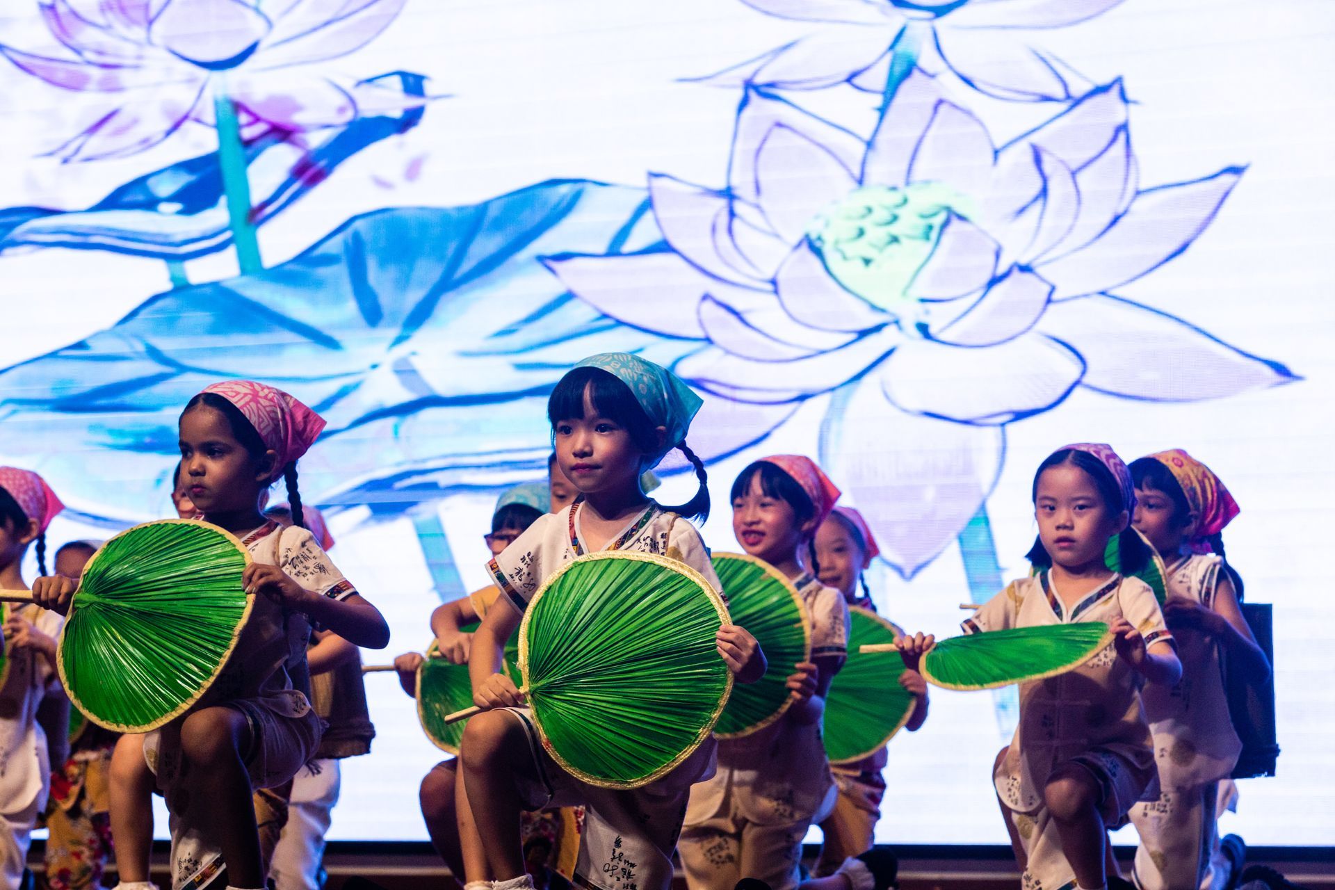 Children performing a dance on stage, holding green fans, with a lotus flower backdrop.