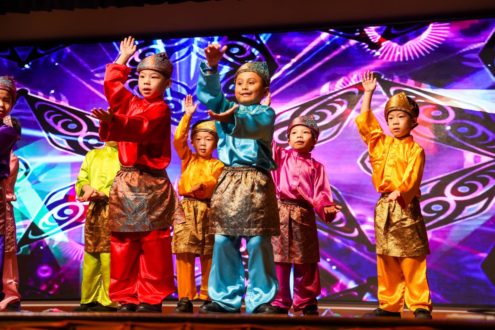 Children in colorful costumes dance on a stage with a bright, patterned backdrop.