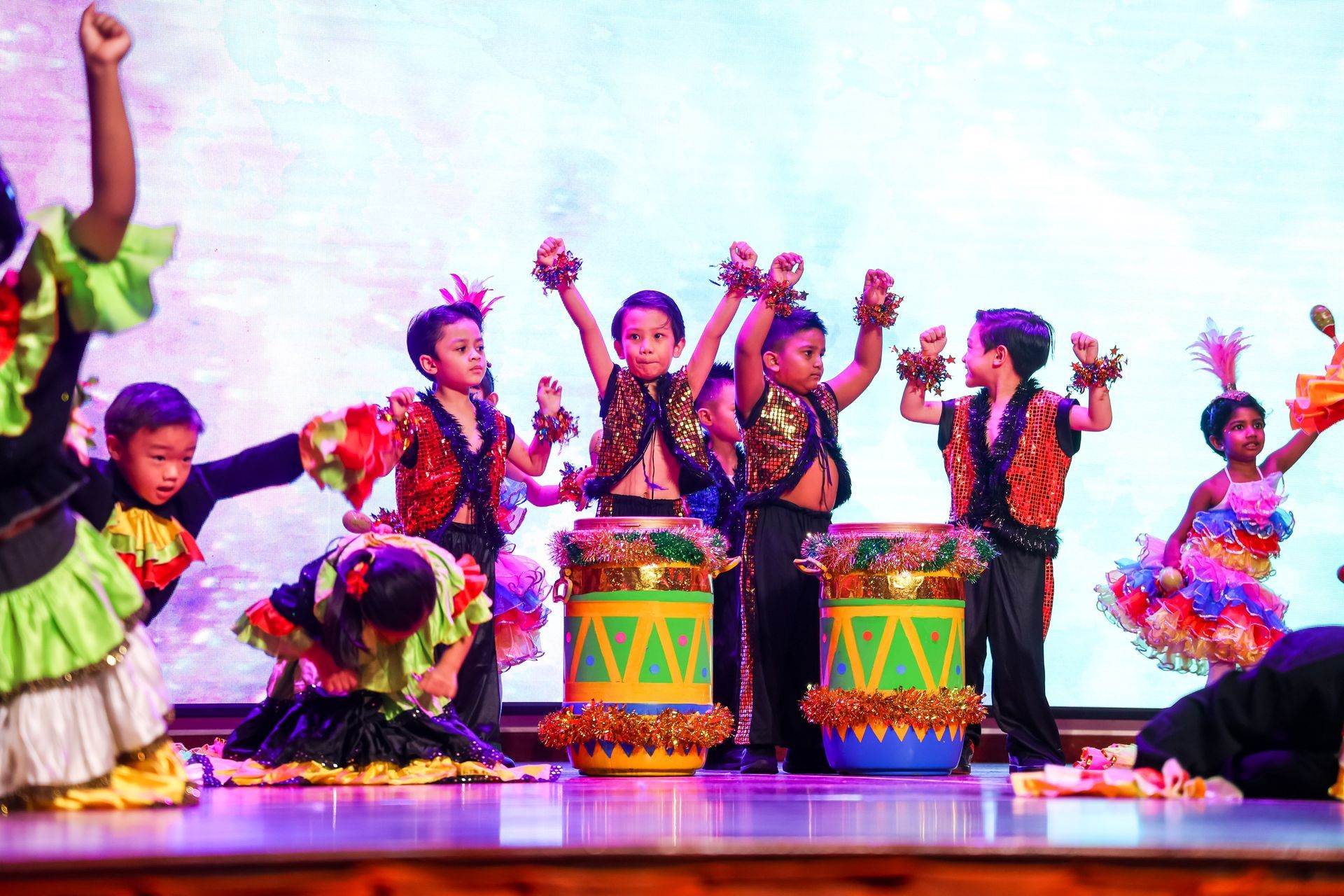 Children in colorful costumes perform on stage, some with drums, raising arms in excitement.