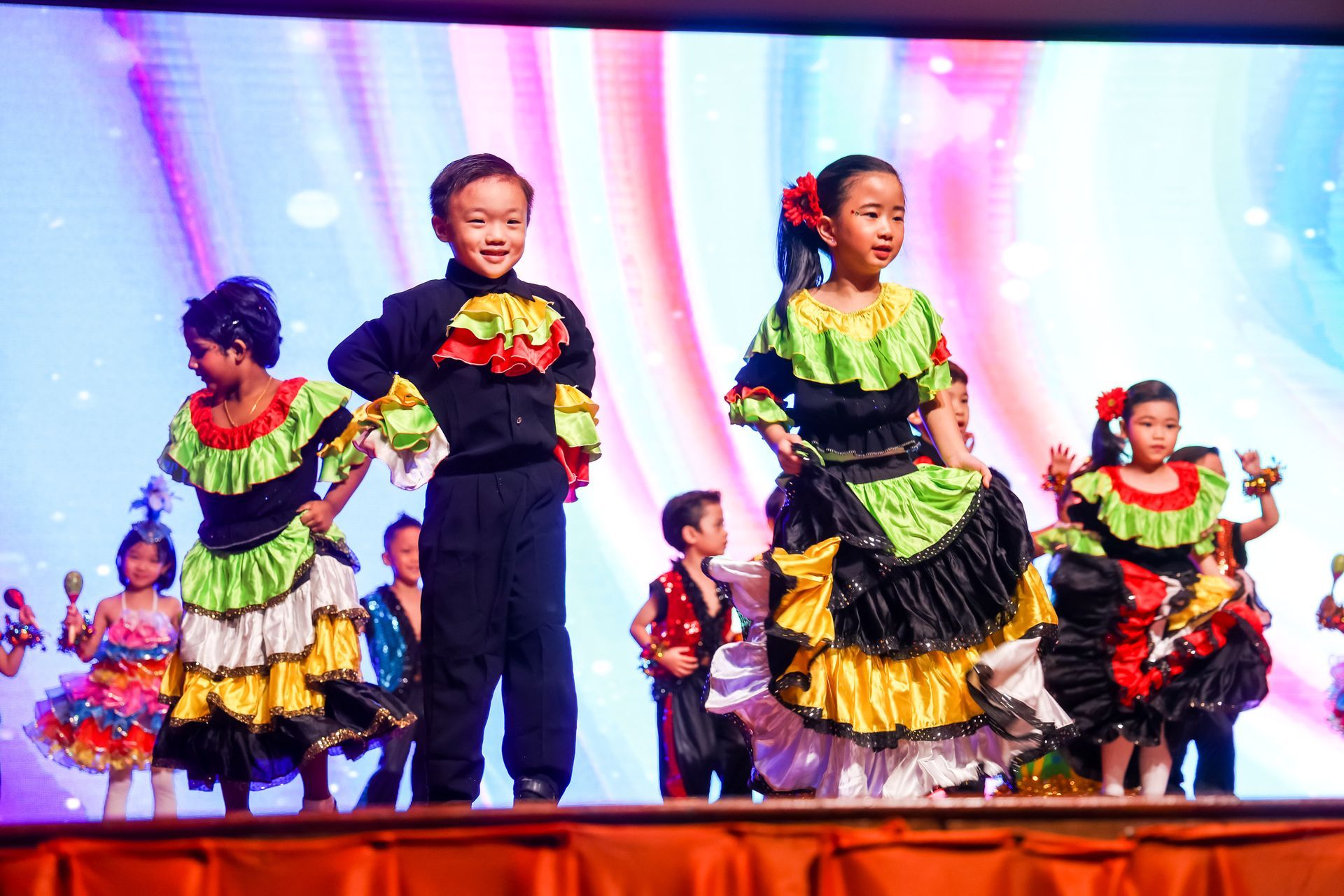 Children in colorful costumes perform on stage, smiling.