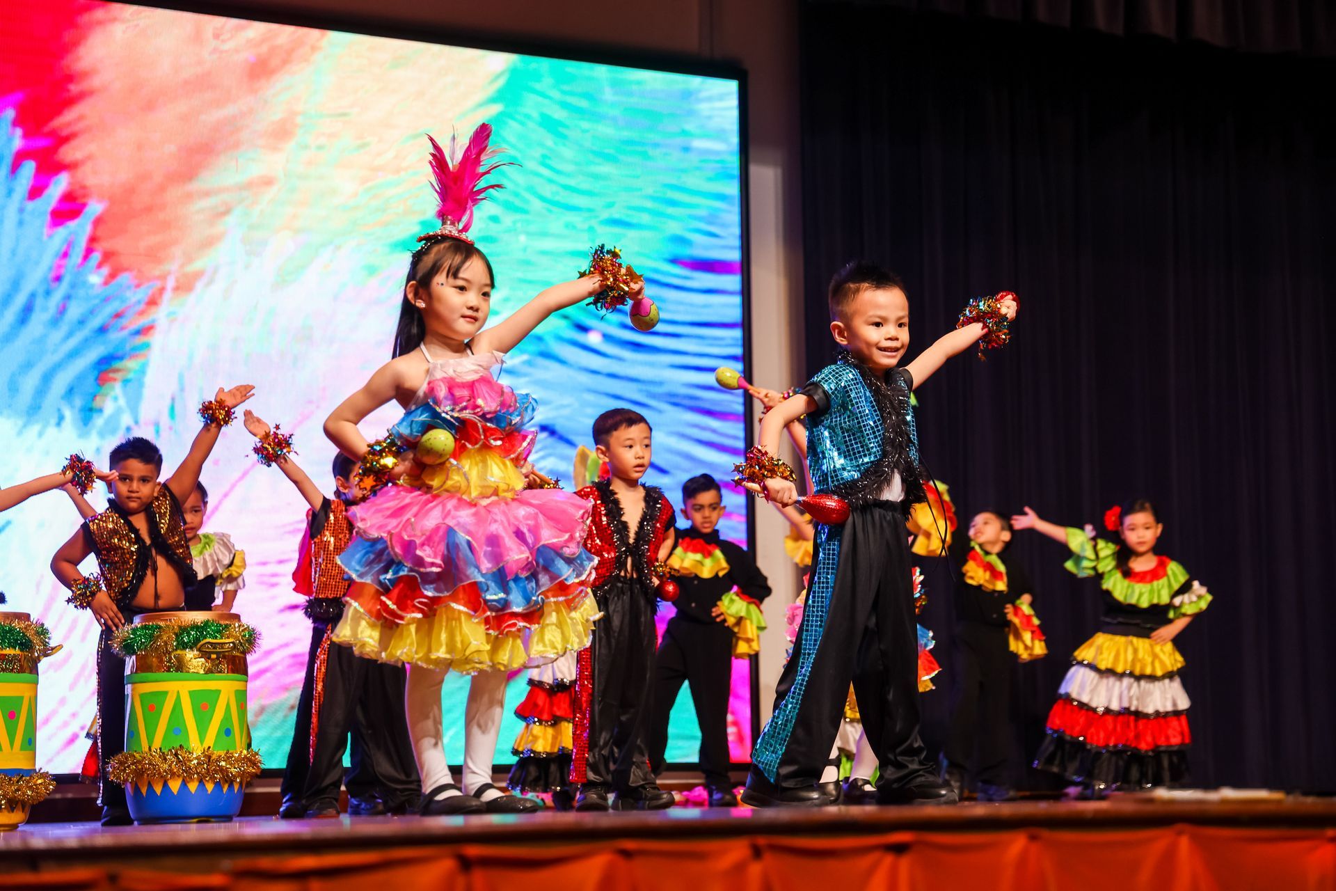Children in colorful costumes perform a dance on stage with a vibrant backdrop.