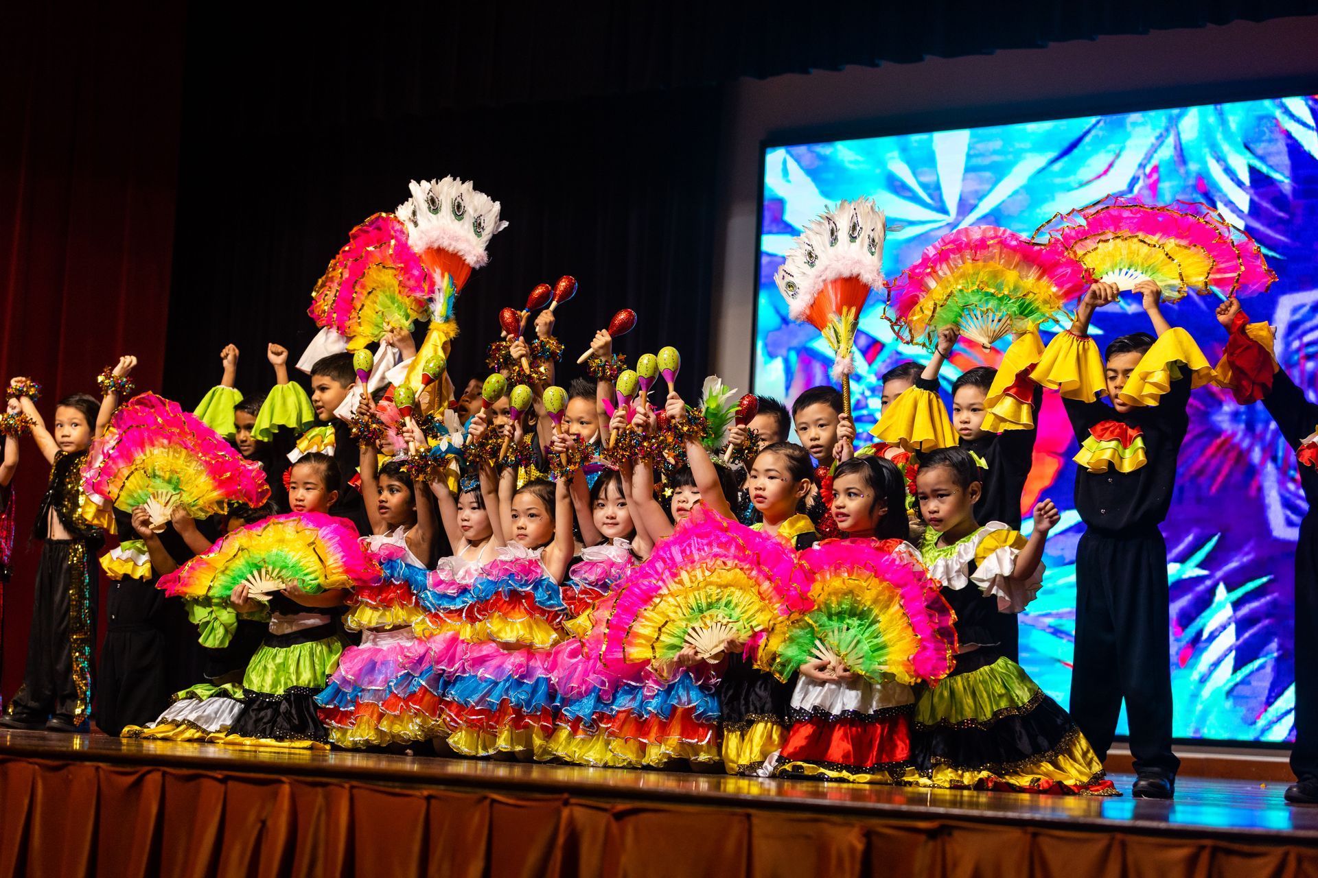 Children in colorful costumes and fans perform on stage.