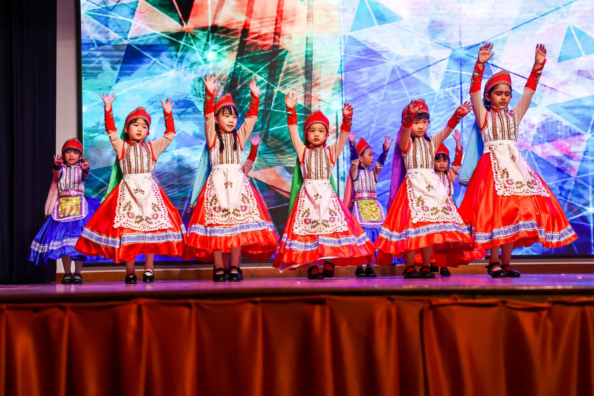 Children in traditional costumes perform on stage, arms raised. Bright colors, backdrop, and audience present.