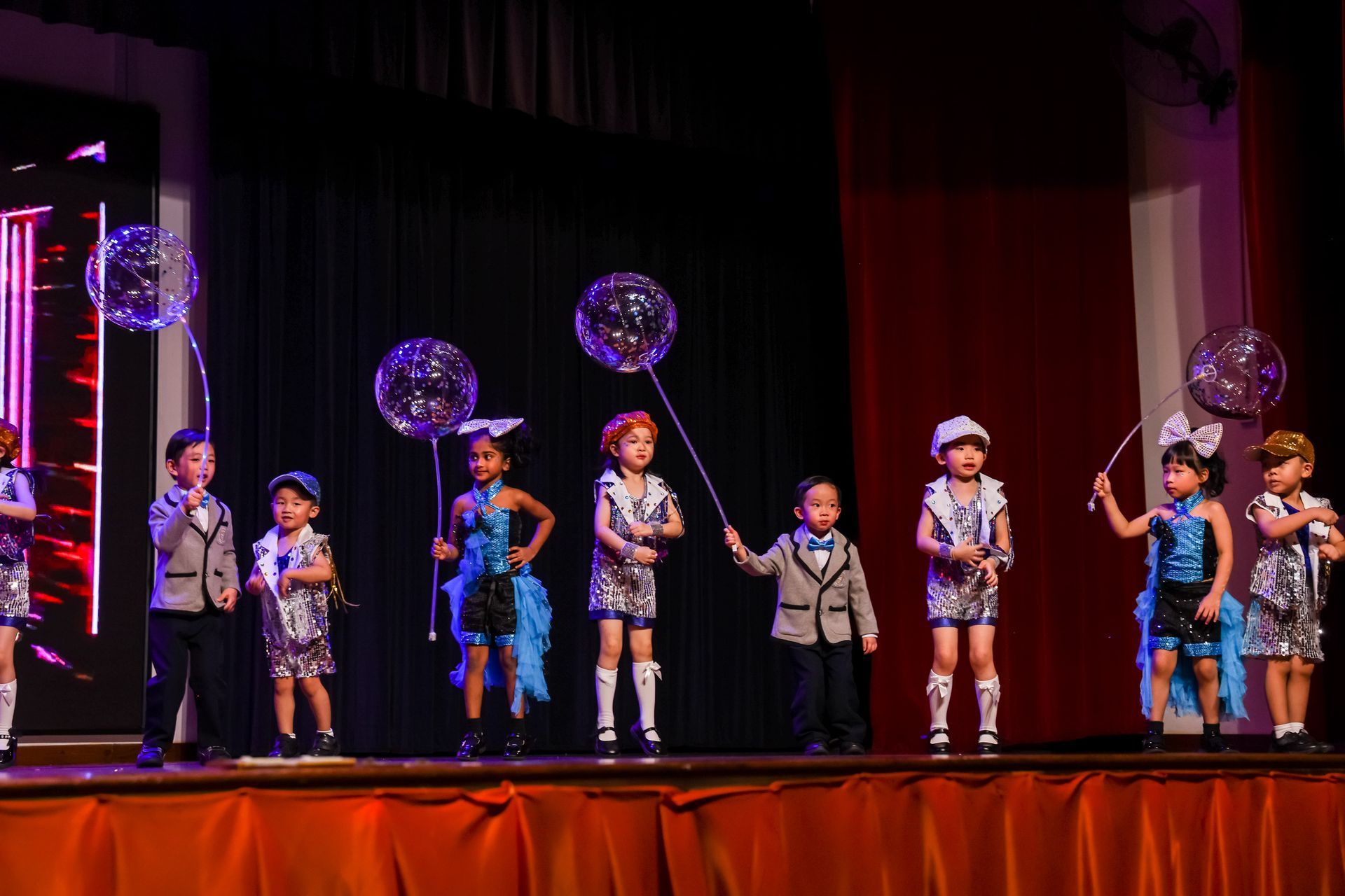 Children on stage in matching outfits, holding balloons. Stage has red and black curtains and lights.