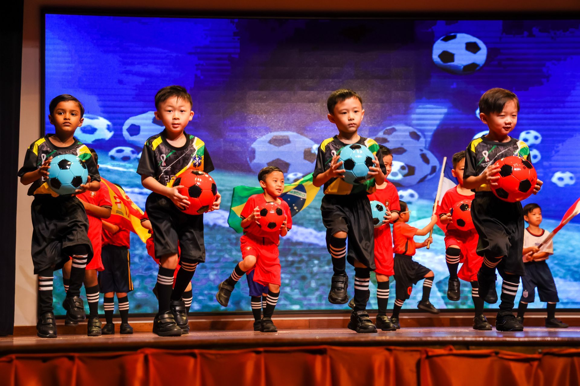 Children performing a soccer-themed dance on a stage, holding soccer balls and flags.