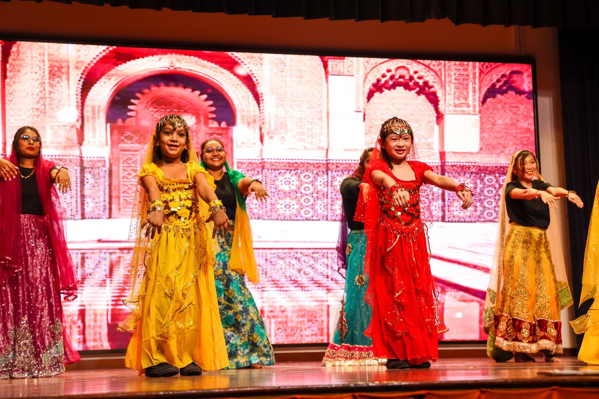 Group of dancers in colorful traditional attire perform on stage, against a backdrop of an ornate archway.