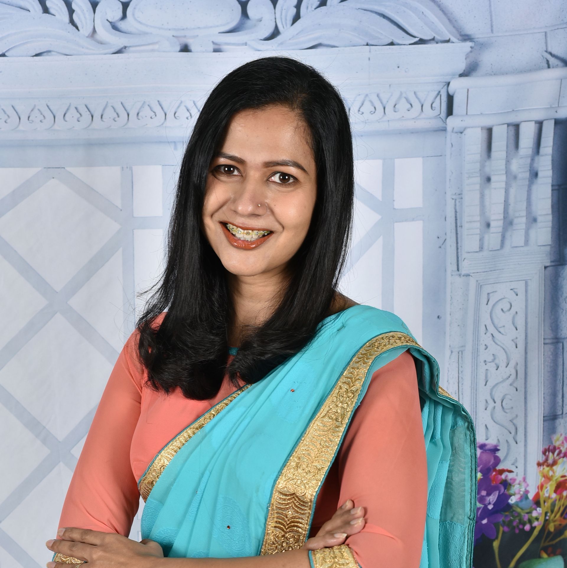 Woman in peach-colored top and turquoise sari smiling, arms crossed, set against a decorated backdrop.