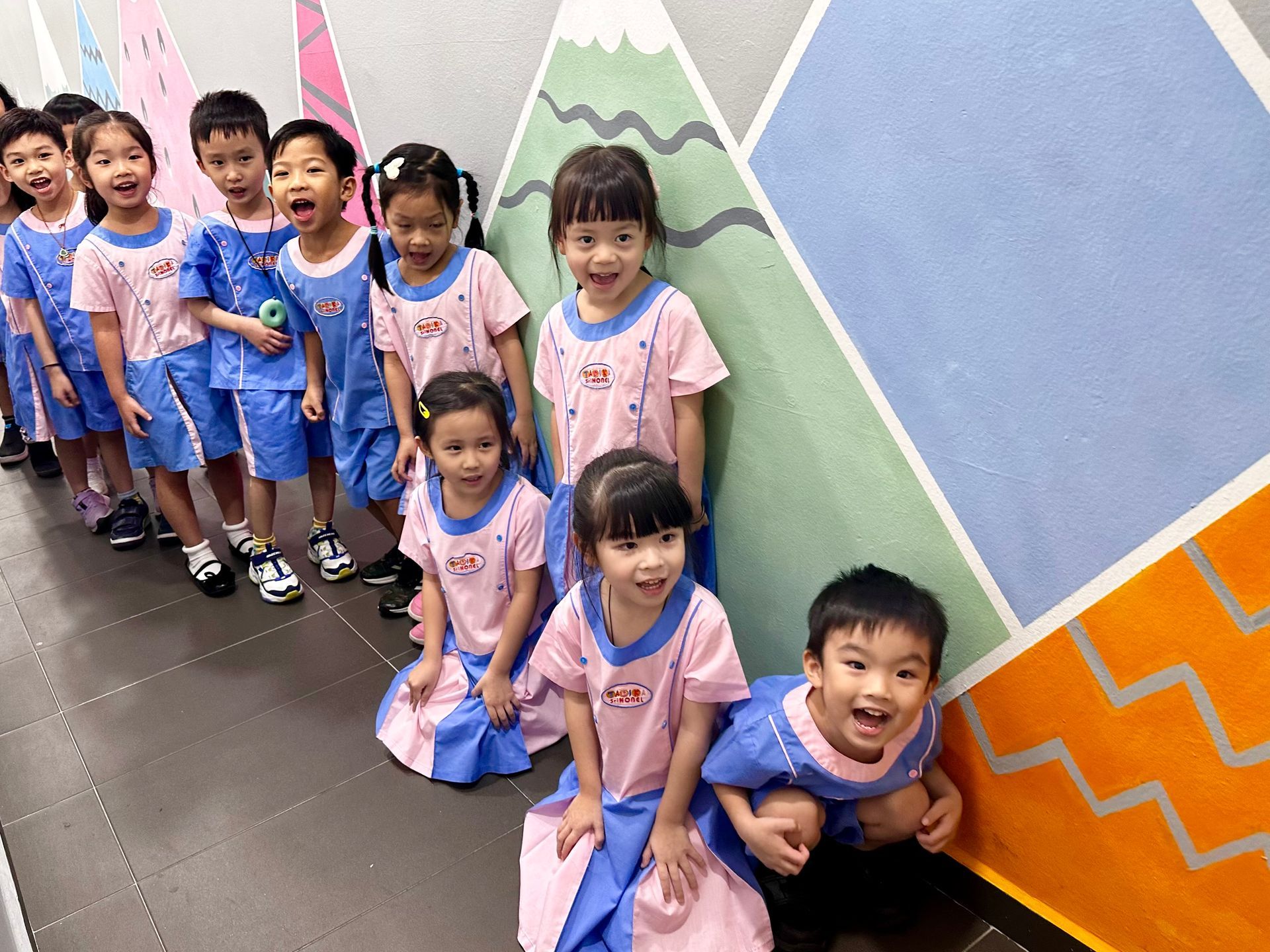 Children in matching uniforms lined up by a colorful wall, some smiling and looking at the camera.