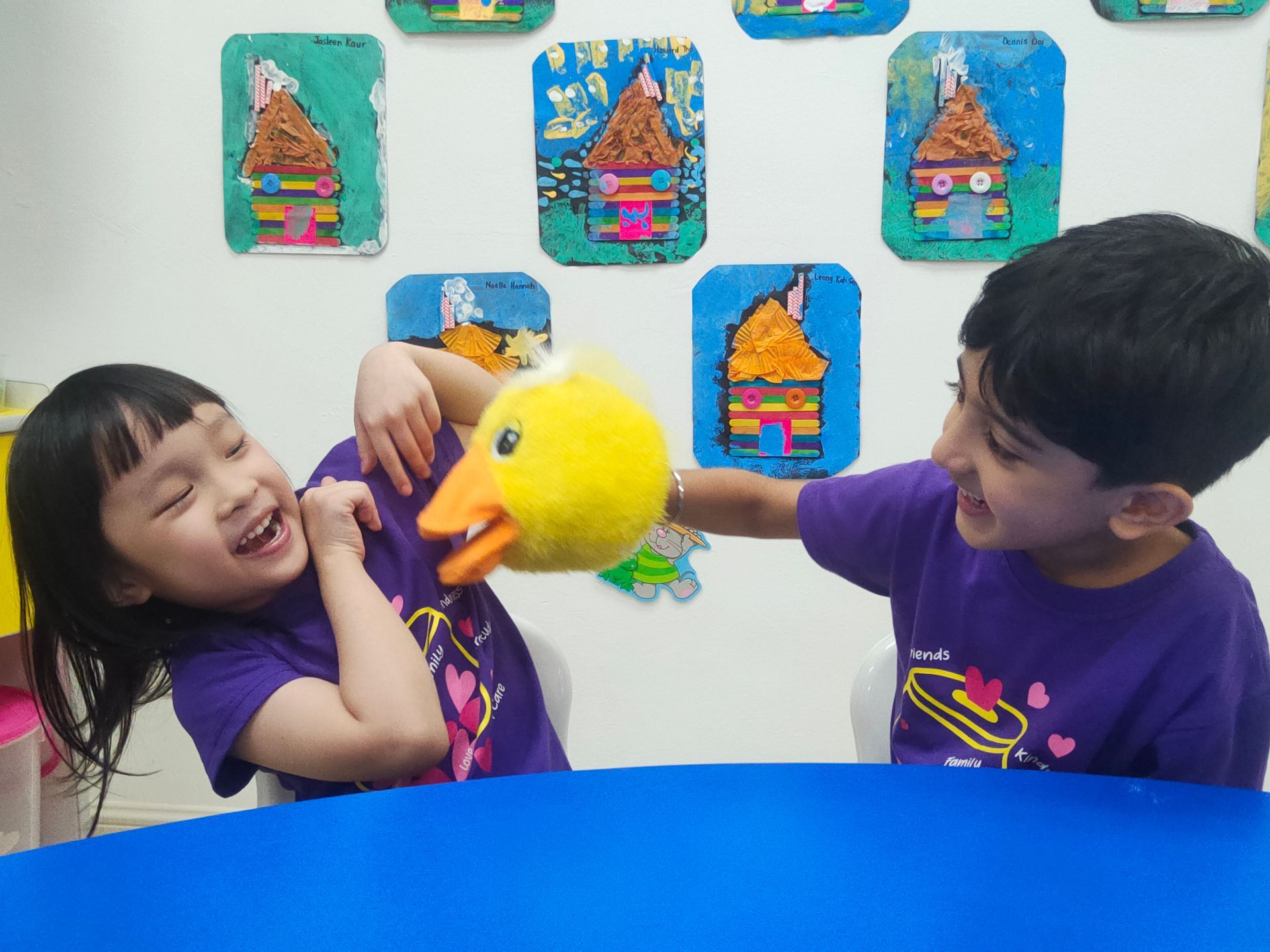 Two children laughing as one playfully touches a yellow duck puppet in front of a white wall decorated with artwork.