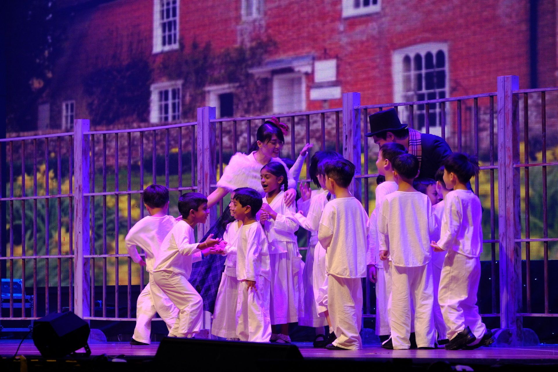 Children in white costumes perform on stage, with a fence and a house backdrop.
