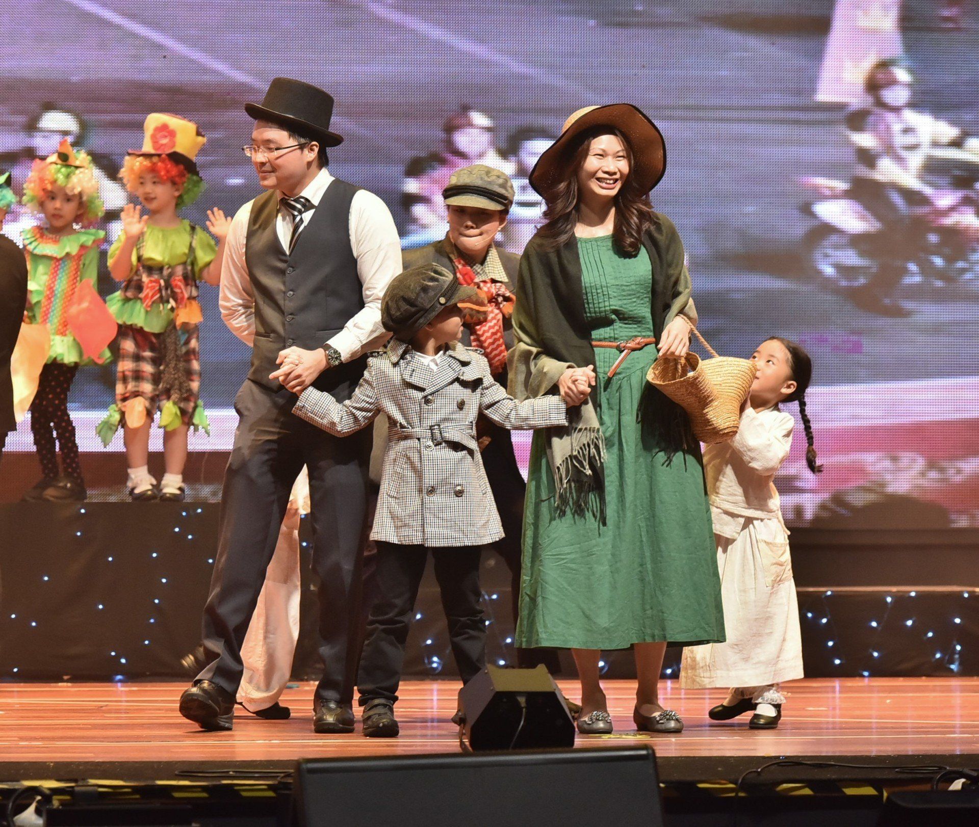 People in costumes on a stage; man in a top hat, woman in a dress, children in various outfits.