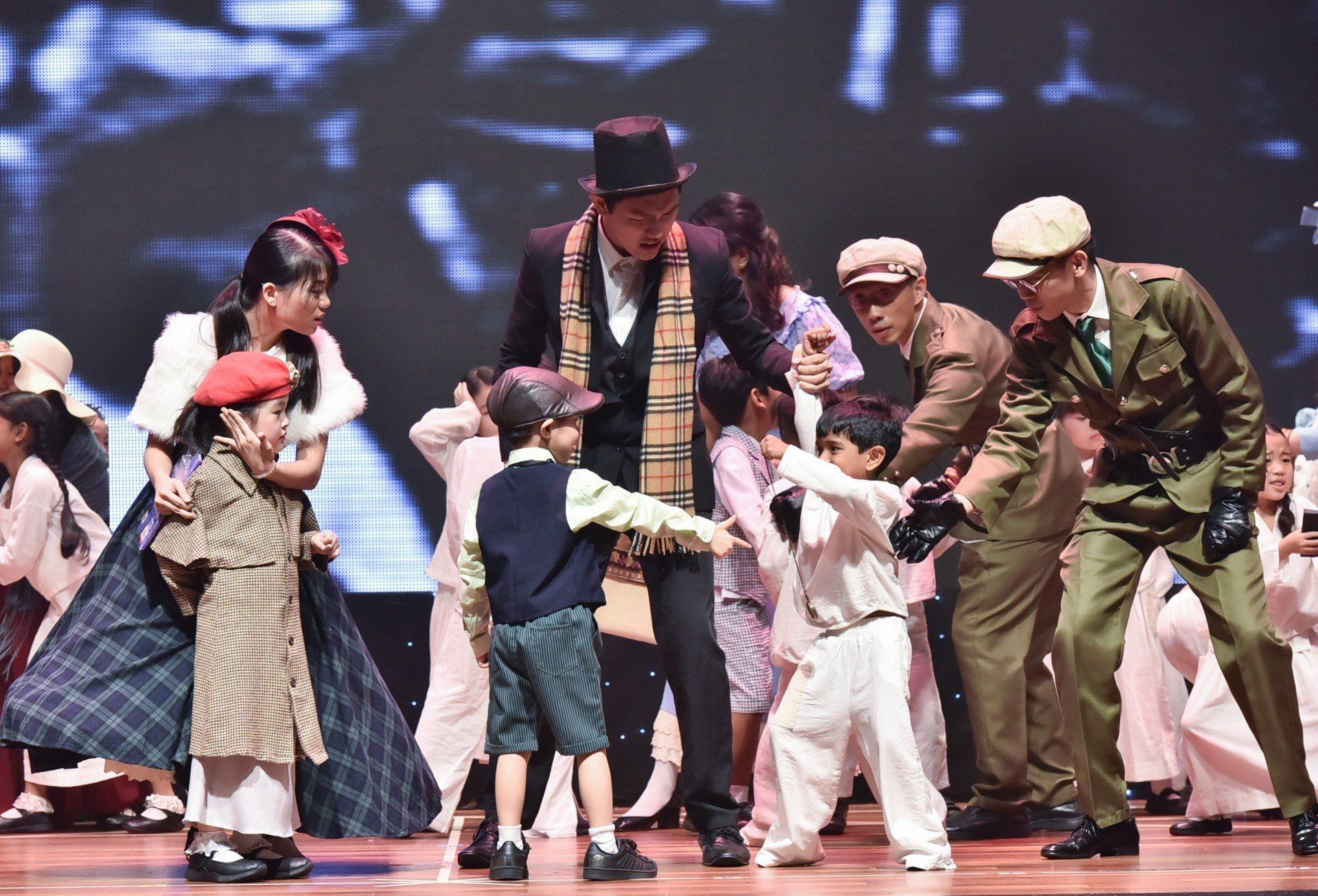 Stage performance: Actors in period costumes, children interacting with a man in a top hat, against a blurred backdrop.