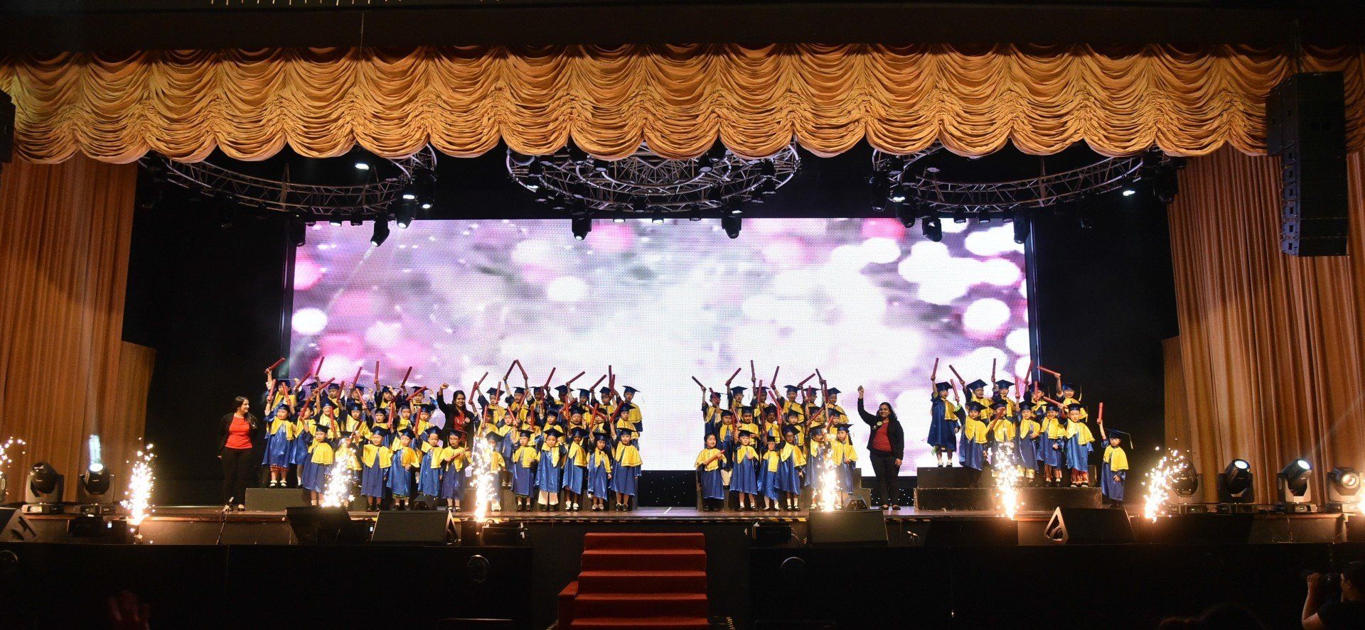 A group of people wearing blue and yellow uniforms on a stage with a screen, celebrating with arms raised.