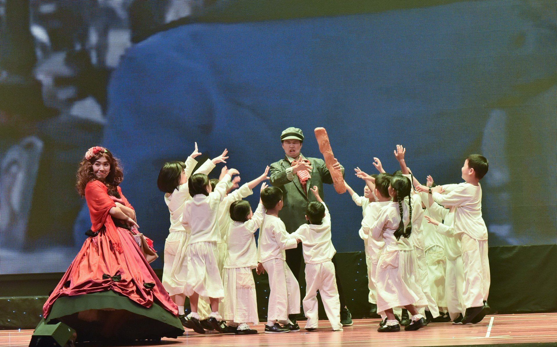 A stage performance: actor in military uniform surrounded by children in white, a woman in a red dress on the side.