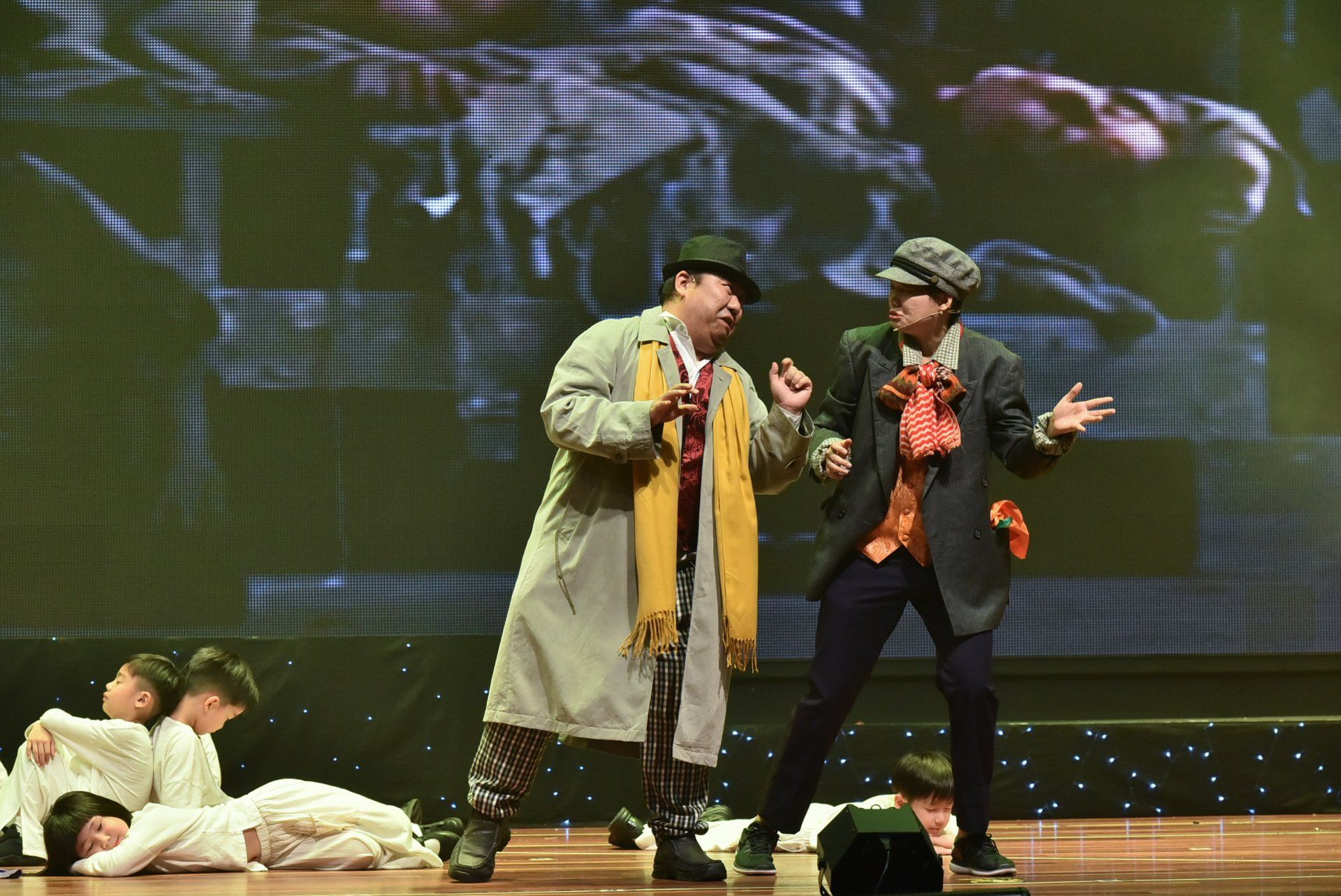 Two men on stage in period costumes perform, children in white lying down.