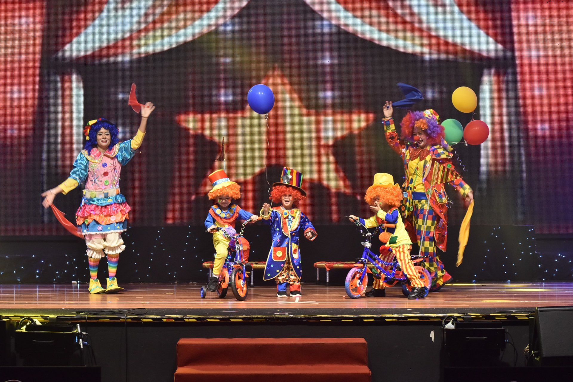 Clowns performing on stage. Costumes are colorful. Balloons and unicycles present. Backdrop features a large star.