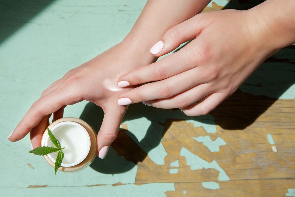 A woman is applying cream to her hands next to a jar of cream.