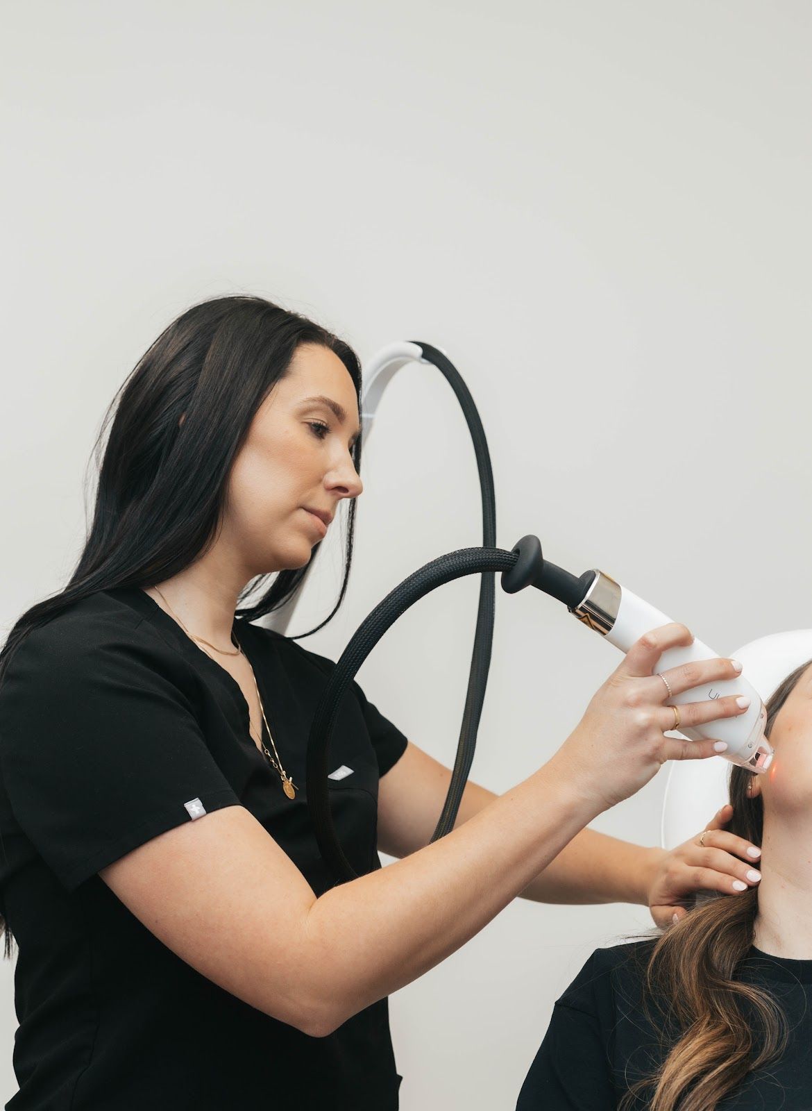 Woman performs laser treatment on another woman's face in a medical setting.