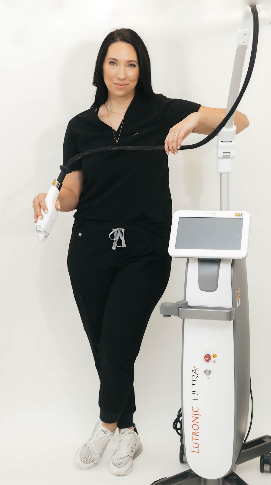 Woman in black scrubs with a medical device, standing in front of a white background.