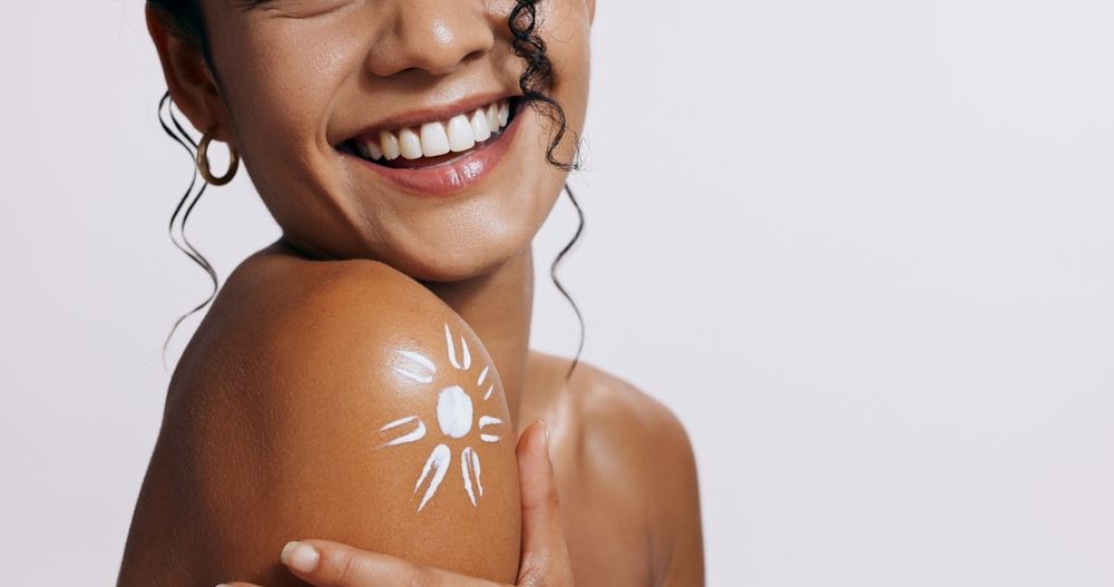 A woman is smiling and applying sunscreen to her shoulder.