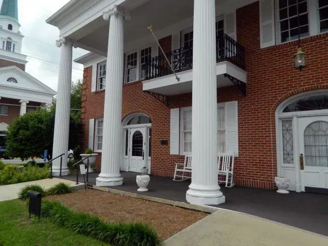 Brick building with white columns and balconies, viewed from a landscaped front walkway.