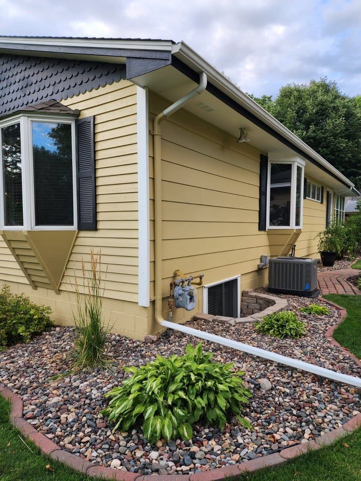 A yellow house with black shutters is surrounded by rocks and plants.