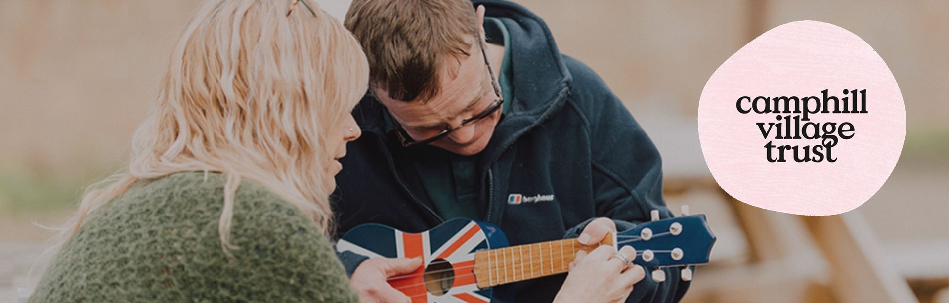 Two people playing a ukulele outdoors. One wears a green sweater, the other a navy jacket. A logo in a pink circle is in the background.