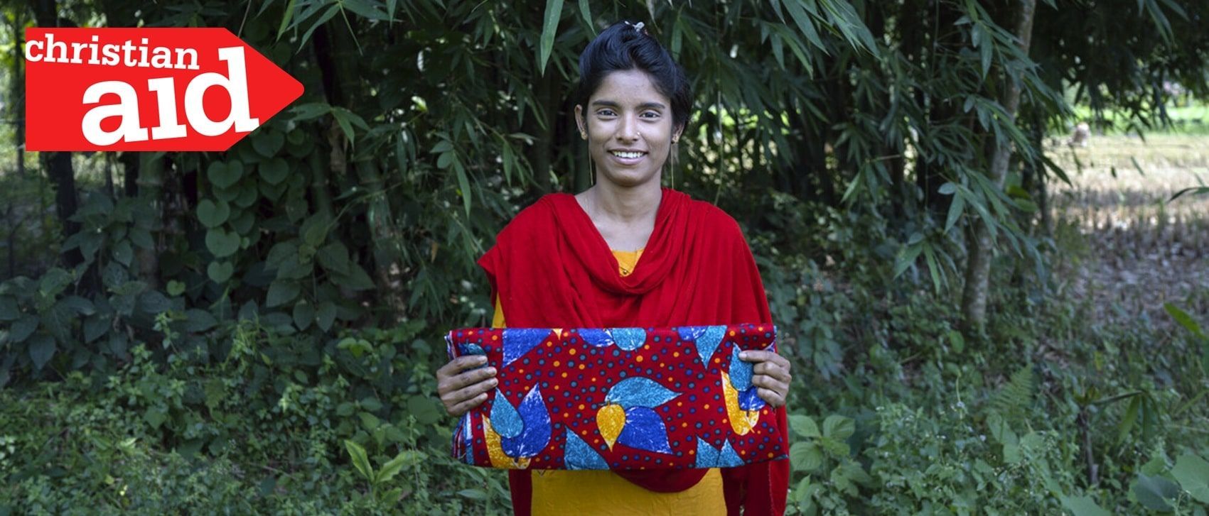 Woman holding patterned blanket, smiling. Red Christian Aid logo in top left. Outdoors, greenery background.