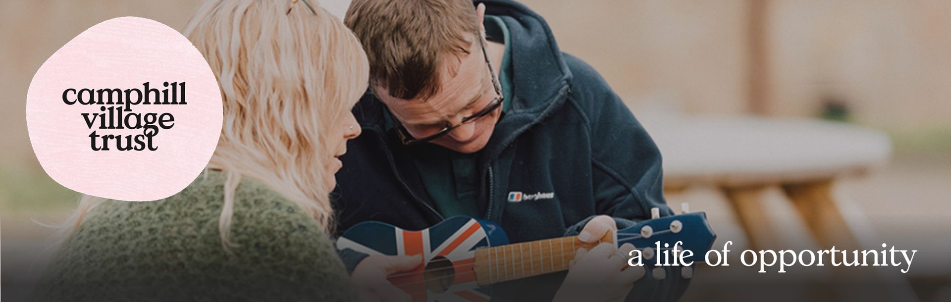 A person plays a guitar while another looks on. Campill Village Trust logo. 