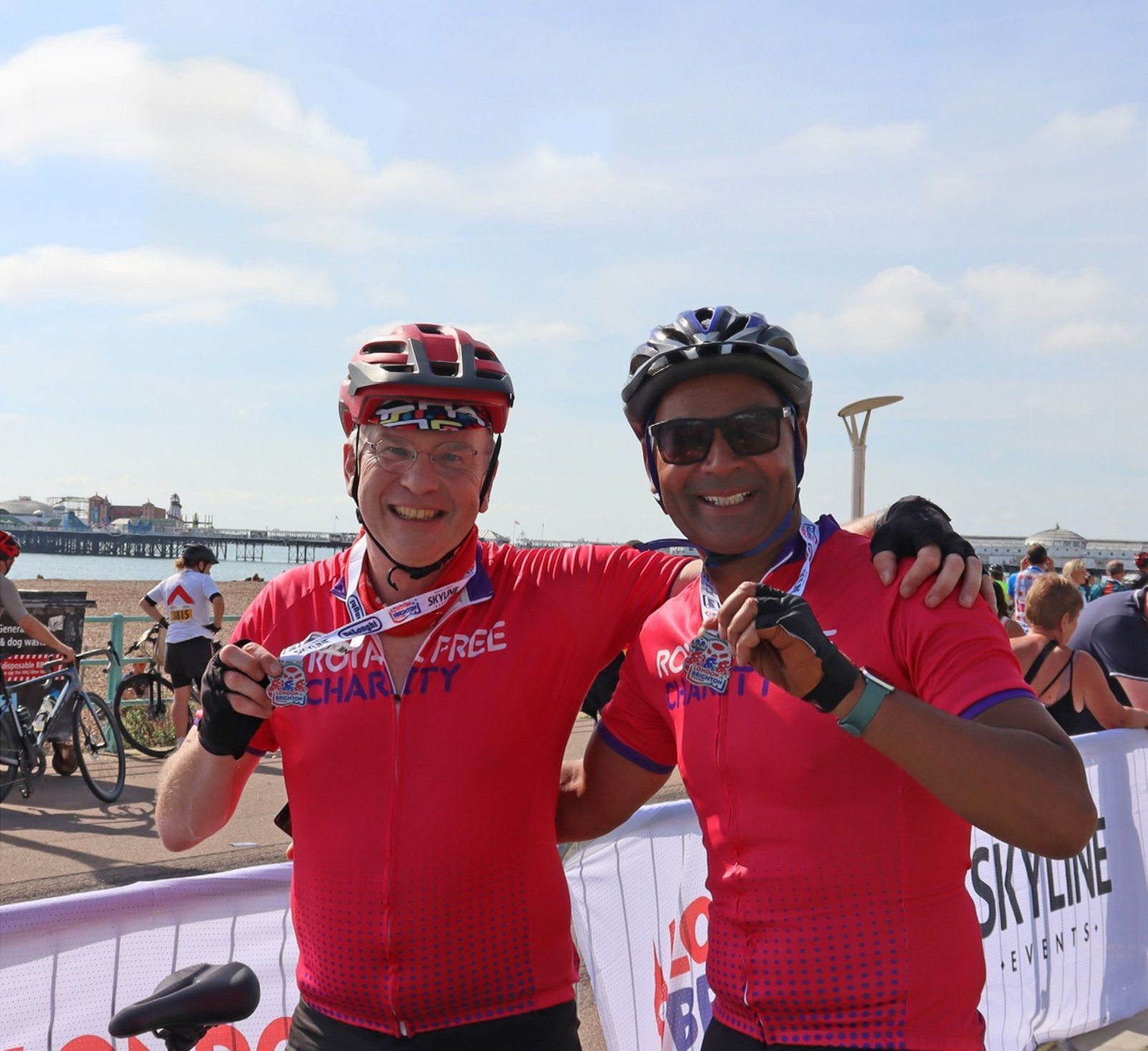 Two cyclists in red shirts and helmets, medals around necks, arms around each other, smiling, near the sea.