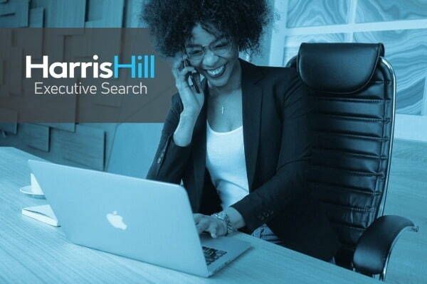 Woman smiles while on the phone and works on laptop; HarrisHill Executive Search logo is visible.