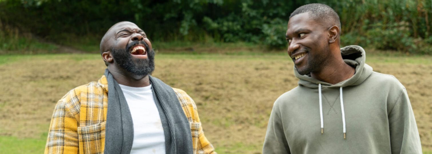 Two men laughing outside. One wears a flannel shirt, the other a hooded sweatshirt.