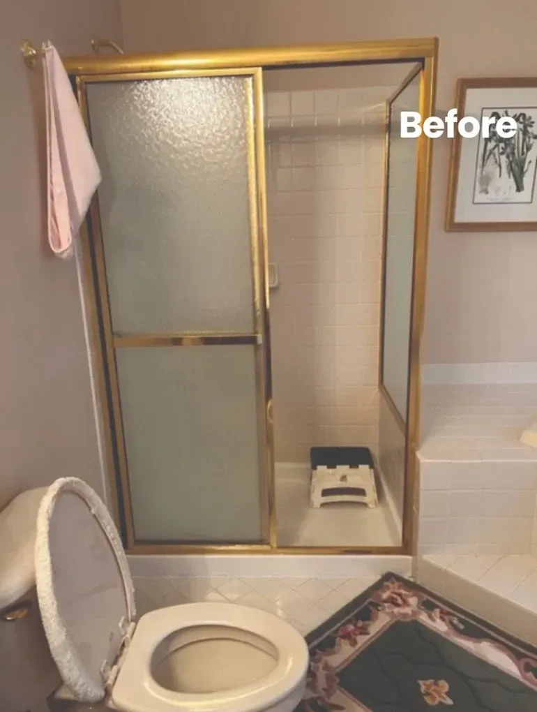 Bathroom with gold-framed sliding shower door and toilet. Before renovation, step stool in shower. Pink towel.