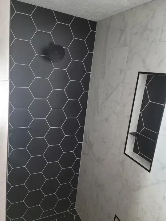 Black hexagonal tiled shower wall with a black shower head, next to a textured grey wall with a recessed shelf.