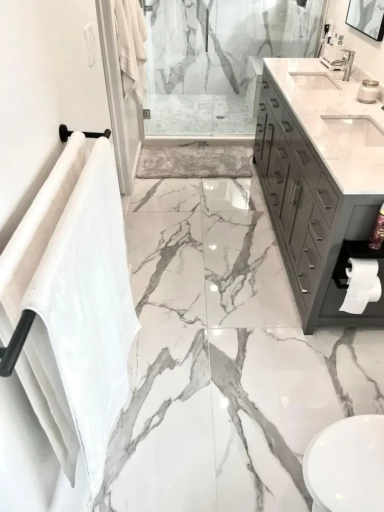 Bathroom with marble-patterned tile, double vanity, glass shower, and white towels on a black towel rack.
