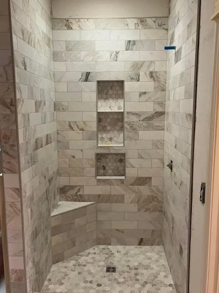Shower tiled in neutral colors with built-in shelves and corner seat; the floor has small hexagonal tiles.