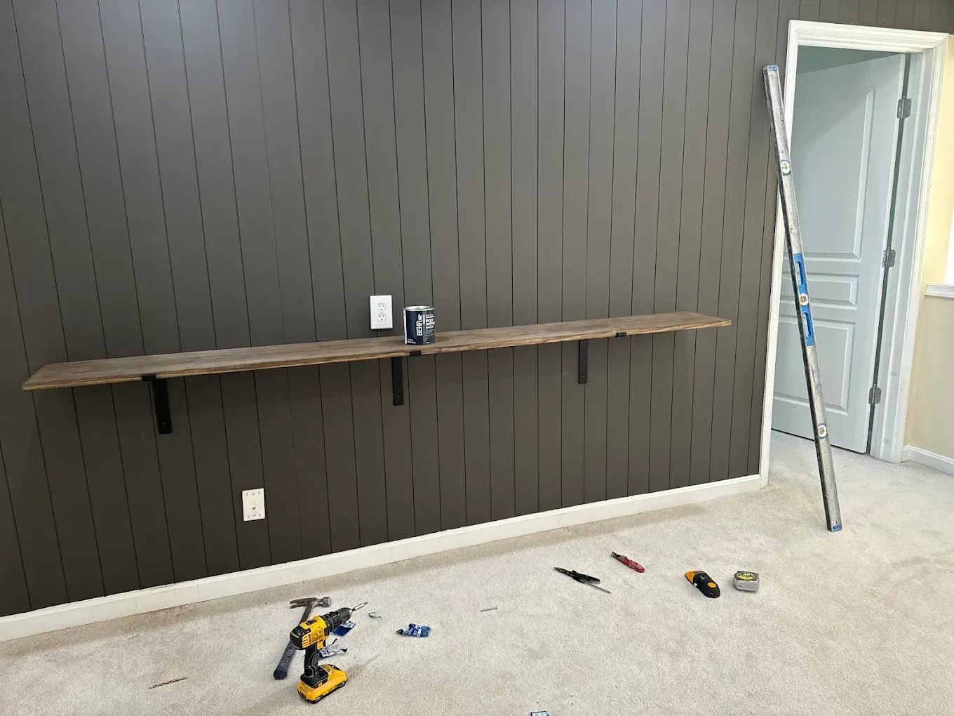 A long, wooden shelf mounted on a dark gray panel wall. Tools and parts are on the carpeted floor below.