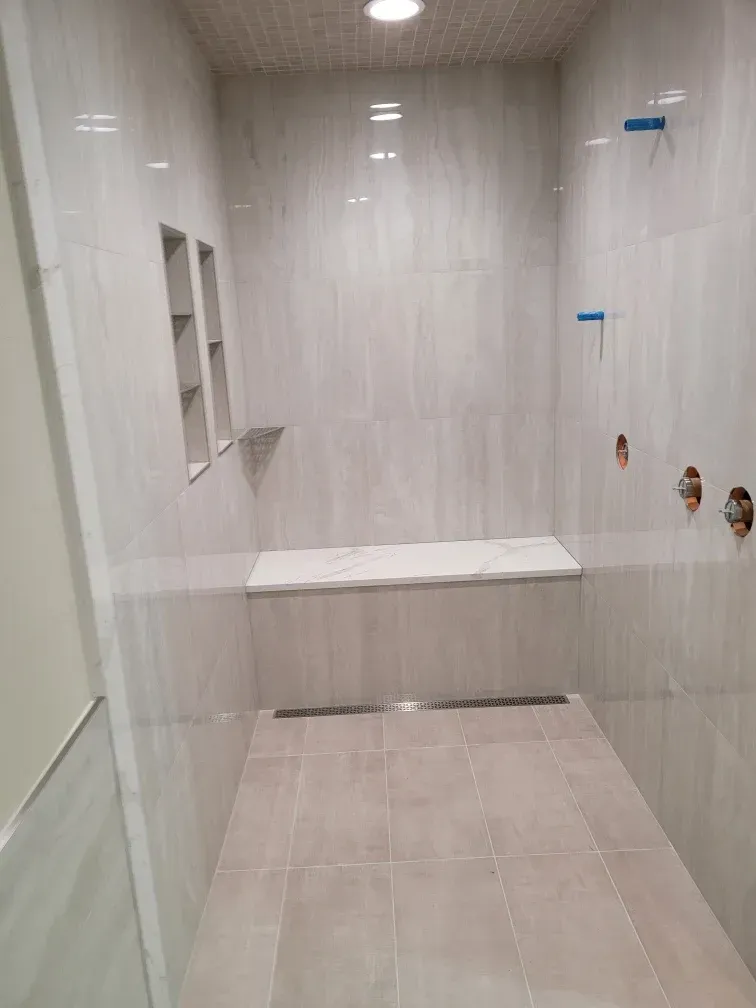 Spacious, tiled shower stall with a built-in bench, recessed shelves, and linear drain. Walls are light grey.
