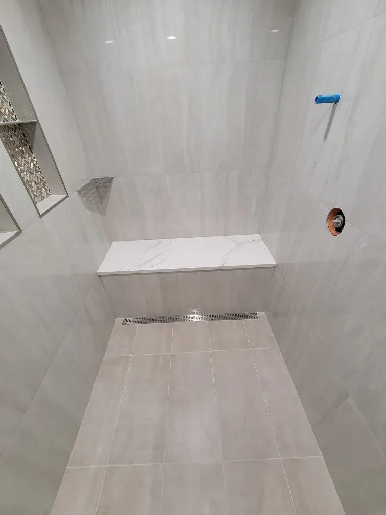 A newly tiled shower stall with a white bench seat, recessed shelves, and linear drain.