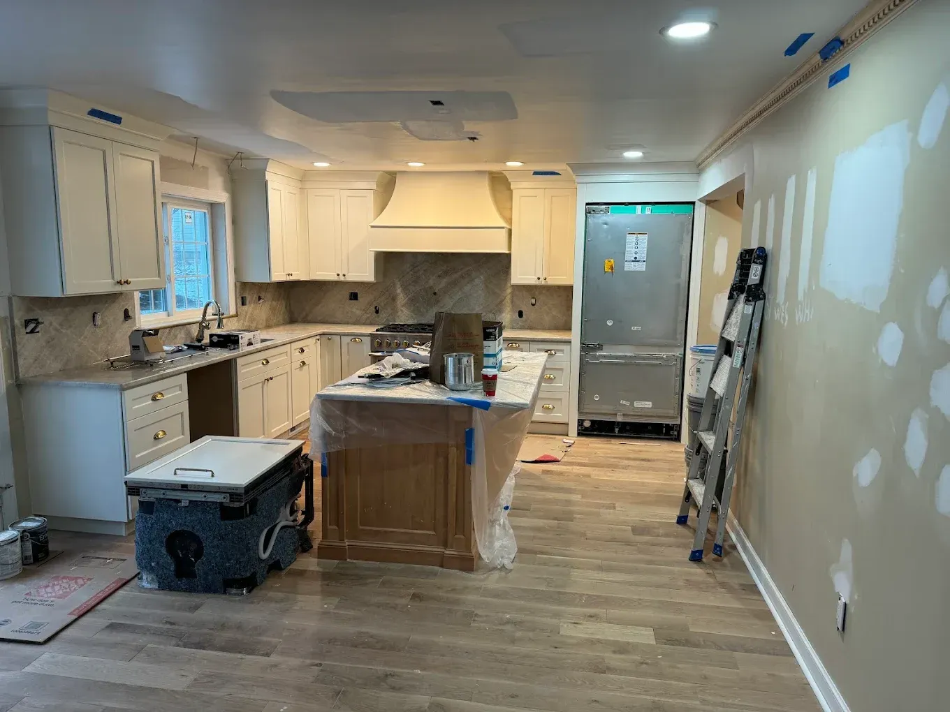 Kitchen renovation in progress: cream cabinets, island, oven hood, partially drywalled walls, and a ladder.