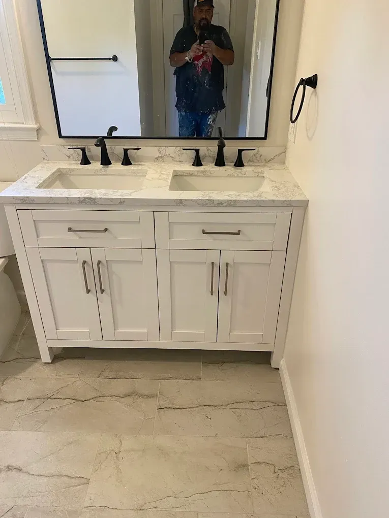 White bathroom vanity with double sinks, black fixtures, and a marble countertop. A person is reflected in the mirror.