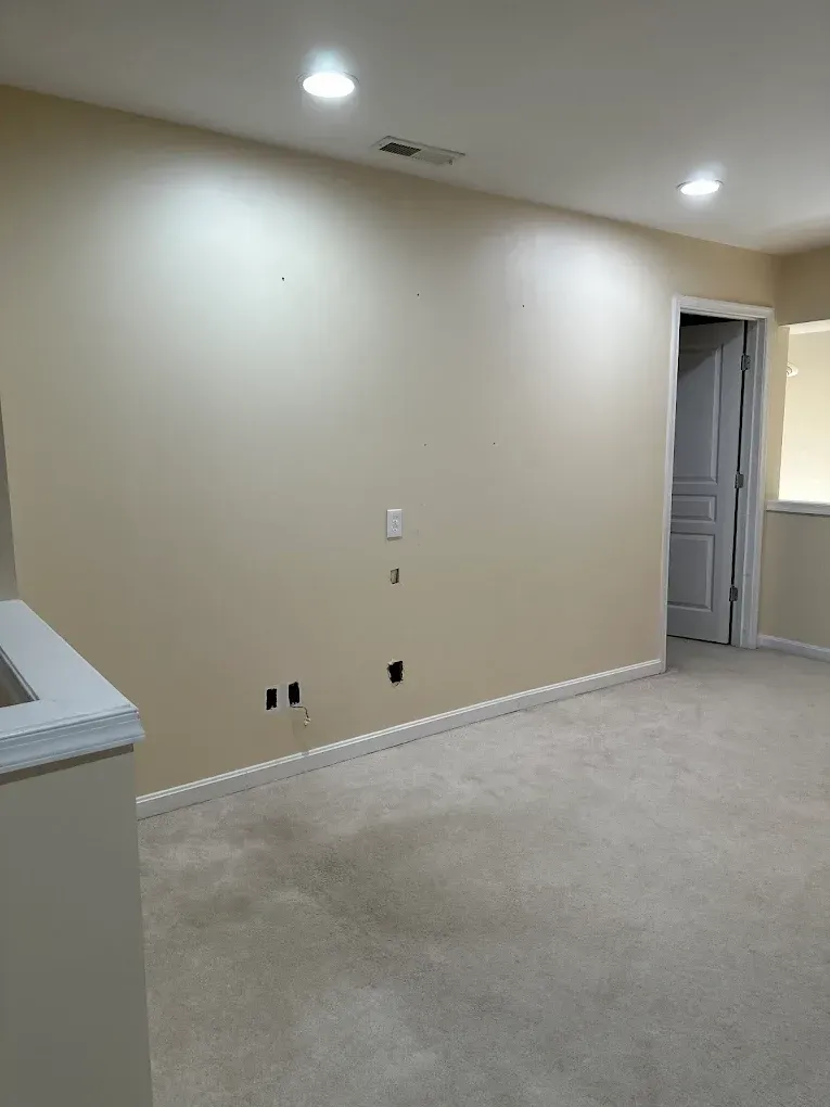 Empty room with beige walls, carpet, and recessed lighting. A door and wiring outlets are visible.