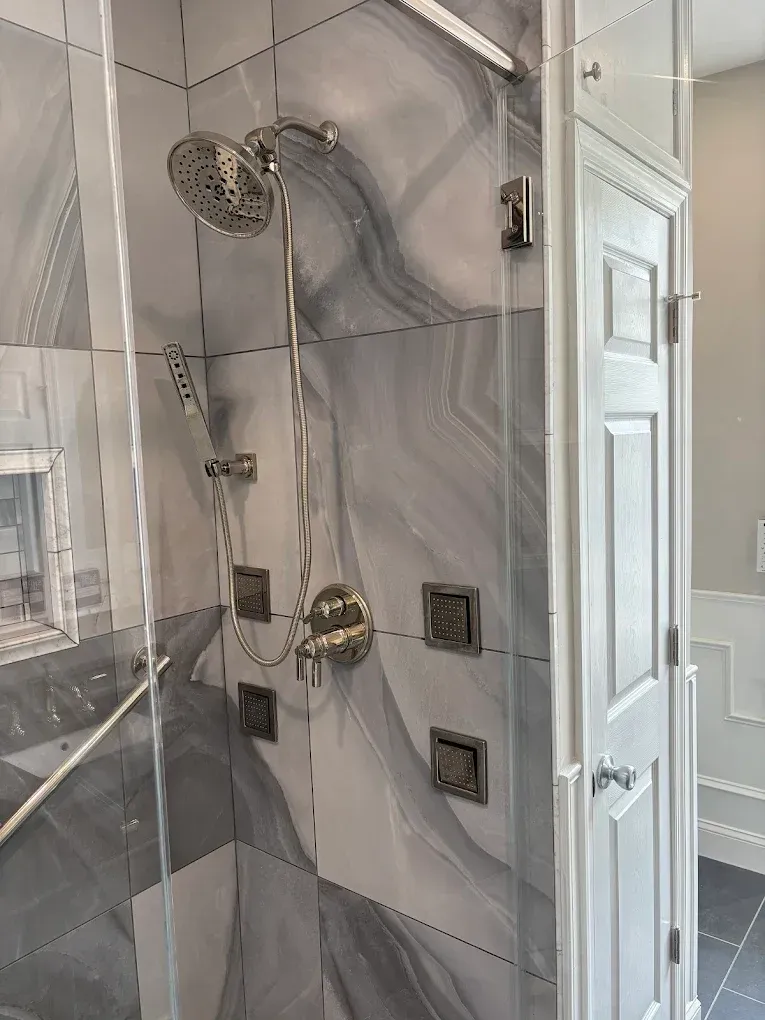 Shower with gray and white marble tiles, glass doors, and multiple showerheads and jets.