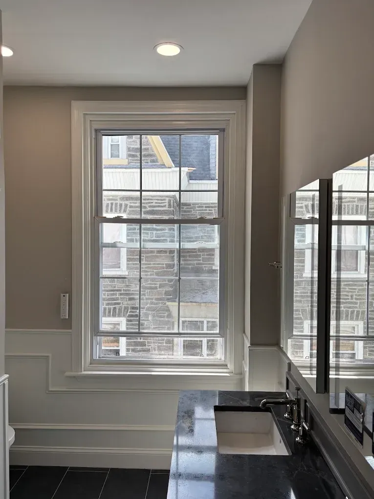 Bathroom with a window looking out to a stone building. White trim, black countertop, and gray walls.