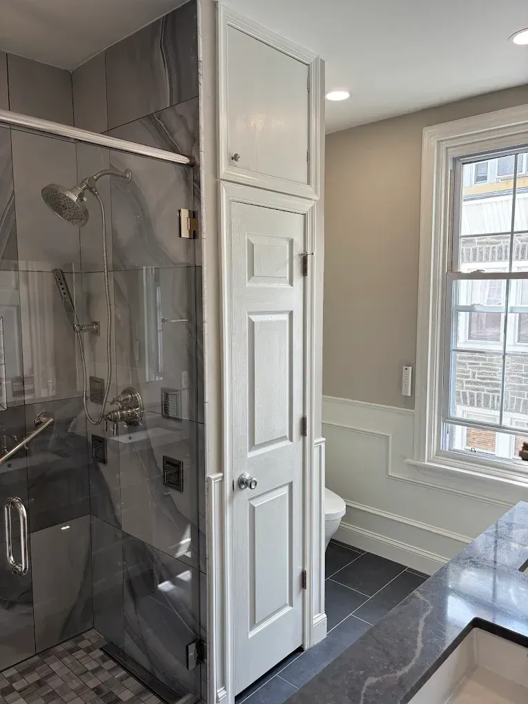 Bathroom with glass shower, tall white storage cabinet, toilet, and window. Gray and white color scheme.